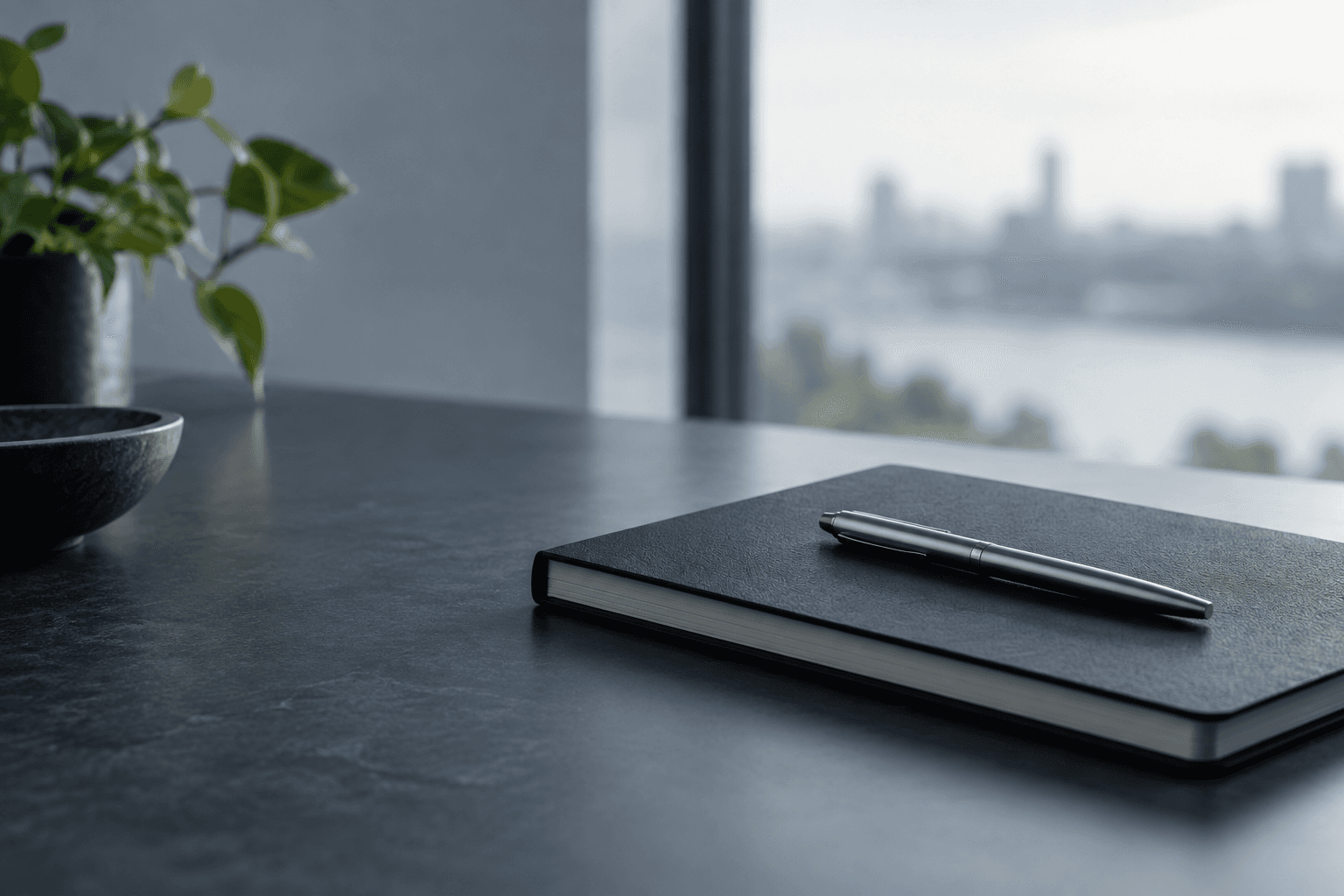 A closed notebook and a pen resting on a dark desk beside a window